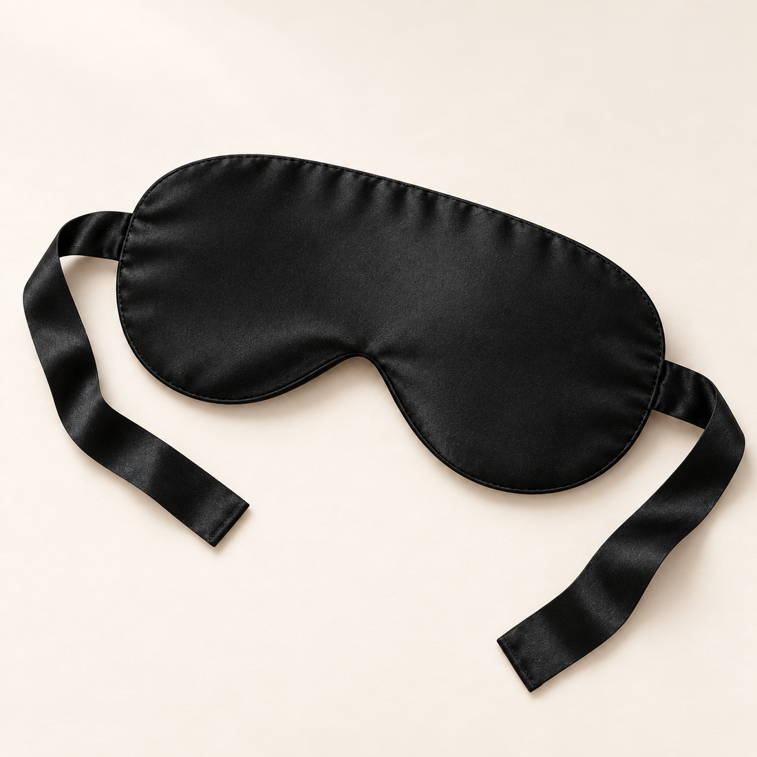 Black satin eye mask with tie straps, shown flat on an ivory background